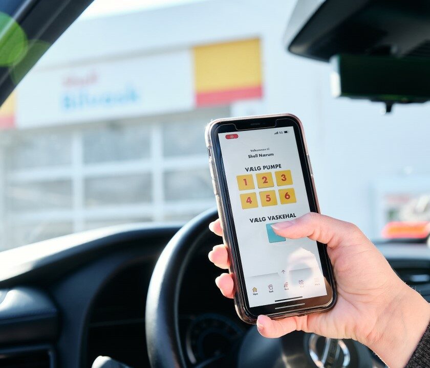 Hånd holder mobiltelefon med Shell Service App i hånden ude foran Shell Bilvask